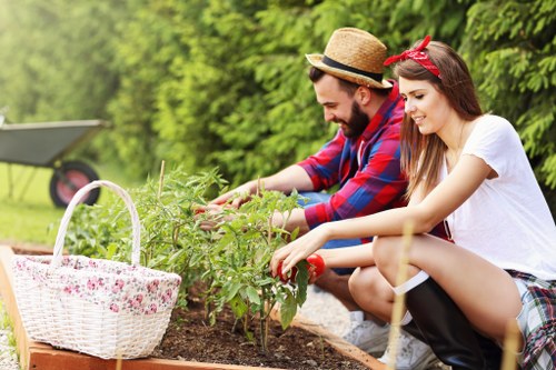 A garden scene in an outdoor space with a landscaped lawn area bordered by a low hedge and surrounded by tall green trees, typical of a garden in Seven Sisters. In the foreground, two individuals—a man wearing a straw hat and a red-and-blue checkered shirt, and a young woman in a white T-shirt with a red headband—are engaged in planting or tending to a small shrub or flowering plant in a raised garden bed with dark, rich soil. To the left, a white basket with a pink floral pattern and a handle rests on the edge of the garden bed. The background showcases a neatly maintained grassy lawn with a garden feature or lawn mower partially visible, and the area is illuminated by natural daylight with clear weather conditions. The scene reflects a well-kept, typical UK-style outdoor garden with attention to plant care and outdoor activity, representative of a professional gardening service provided by Landscapers Seven Sisters.