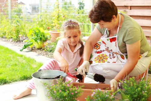 In a well-maintained garden area, a woman and a young girl are engaging in gardening activities together. The woman, wearing an apron with a floral pattern, is kneeling on the ground and appears to be assisting the girl, who is sitting cross-legged on the grass. They are working with a rectangular plant bed that is filled with dark soil, possibly planting or tending to plants. The garden features a lush, green lawn, with various flowering plants and shrubs bordering the garden bed. In the background, there is a wooden garden shed, along with a vegetable patch or flower bed enclosed by a wire fence. The scene is lit by natural daylight, suggesting a clear weather day, and the overall environment indicates a spacious outdoor area typical of gardens in the Seven Sisters region, suitable for gardening and outdoor maintenance services by Landscapers Seven Sisters.