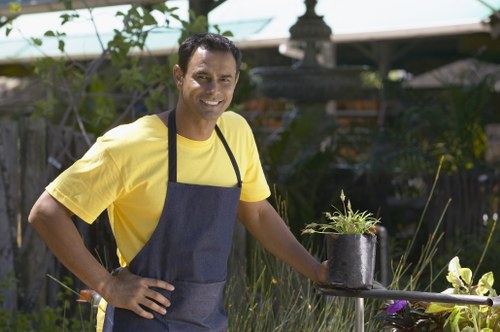 A man wearing a yellow T-shirt and a dark apron stands outdoors in a landscaped garden, smiling at the camera. The garden features a well-maintained lawn with dense, green grass in the foreground, bordered by flower beds and shrubbery. Behind him, there are various plants, including tall grasses and a small potted plant that he is holding, placed on a short table. The background shows additional garden elements such as wooden fencing, a greenhouse, and some potted plants, indicating a neatly designed outdoor space typical of residential gardens in the Seven Sisters area. The environment appears sunny with clear weather, highlighting the natural tones of the plants and the vibrant outdoor setting, aligning with gardening and landscaping services offered by Landscapers Seven Sisters.