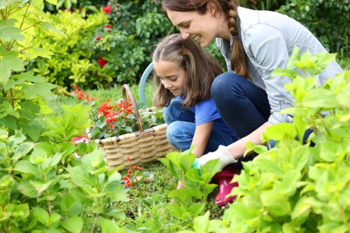 A woman and a young girl are gardening together in a lush backyard, surrounded by vibrant green shrubs, leafy plants, and blooming flowers. The woman is kneeling next to the girl, who is holding a small wicker basket, suggesting they are collecting or planting garden produce. The garden features a well-maintained lawn with dense grass, bordered by a variety of mature hedge plants and flowering bushes, creating a natural and inviting outdoor space. Behind them, a mix of trees and flowering plants provides a colorful and textured backdrop, with natural sunlight illuminating the scene and highlighting the greenery and floral hues. The garden surface appears to include a combination of soil beds and paved pathways, typical of a professionally landscaped garden maintained by a local gardening service such as Landscapers Seven Sisters. This image captures a peaceful moment of outdoor activity, emphasizing the beauty and diversity of a well-kept garden environment in a suburban setting near postal code SE, supporting outdoor maintenance and gardening expertise.