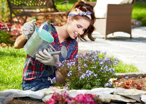 A young woman with light skin and shoulder-length brown hair, wearing a blue and red plaid shirt, grey gardening gloves, and a headband, is kneeling in a well-maintained garden. She is watering a small flowering shrub with light purple blossoms using a metal watering can. The garden features a curved stone border separating a lush, green lawn from flower beds that contain various plants and flowers of different colours, including pink and red blooms. In the background, there is outdoor furniture including a patio table and chairs on a paved area, with a wooden fence and trees suggesting a suburban garden environment typical of the Seven Sisters area. The weather appears sunny and bright, with natural light highlighting the vibrant plants and the woman's cheerful expression. The scene exemplifies typical garden maintenance and planting activities supported by structures and plant arrangements common in domestic outdoor spaces, aligning with professional gardening services provided by Landscapers Seven Sisters and their focus on lawn care, planting, and yard aesthetics in the local postcode area.
