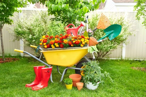 A garden scene featuring a brightly painted yellow wheelbarrow filled with vibrant red and yellow chrysanthemums, positioned on a well-maintained lawn with lush green grass. Surrounding the wheelbarrow are various gardening tools and accessories, including red rain boots, a small terracotta pot, a white pot with a green leafy plant, and other smaller plant containers. In the background, a white picket fence encloses the garden area, with mature trees and flowering shrubs creating a natural backdrop. The scene appears to be in a residential outdoor space, possibly in Seven Sisters, and suggests active gardening or planting activities during daylight hours, with soft natural lighting and a clear, pleasant weather condition.