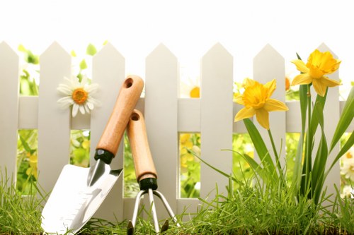 A close-up view of a well-maintained garden border featuring a white picket fence with evenly spaced vertical slats, set against a background of blurred greenery. In the foreground, lush green grass and dense planting are visible, with a variety of blooming flowers including bright yellow daffodils and white daisies with yellow centers. The garden scene appears to be outdoors in natural daylight, suggesting a sunny or partly cloudy day, with the plants flourishing in a temperate climate typical of the Seven Sisters area. Gardening tools such as a small hand trowel with a wooden handle and a pair of pruning shears are placed on the grass, indicating recent or ongoing gardening activity. The overall setting reflects a carefully curated and landscaped outdoor space suitable for garden maintenance services offered by Landscapers Seven Sisters, emphasizing flower bed creation and general gardening work in a suburban or residential environment near SE postcodes.