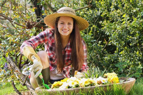 A young woman with long brown hair, smiling and wearing a straw sunhat, plaid shirt, gardening gloves, and dark trousers, is kneeling in a lush garden. She is actively planting or tending to a flower bed filled with vibrant yellow and purple blooms, surrounded by green foliage and shrubs. The garden features a well-maintained lawn area in the foreground, with a backdrop of dense trees and hedges, indicating a landscaped outdoor space typical of residential gardens in the Seven Sisters area. The natural lighting suggests a bright, sunny day, enhancing the vivid colours of the flowers and foliage. This scene depicts outdoor gardening activity, showcasing careful planting and garden maintenance, which aligns with professional gardening and landscaping services provided by Landscapers Seven Sisters, emphasizing their expertise in garden design, planting, and outdoor beautification control.