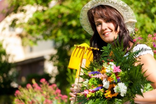 A woman with shoulder-length dark brown hair, wearing a wide-brimmed straw hat and a white top, standing in a lush garden during daytime, holding a large bouquet of colourful flowers and greenery. Behind her, there is a well-maintained lawn with dense grass, a pathway edged with small flower beds, and a background of mature trees with green foliage. The garden features neatly trimmed hedges, blooming flower patches with pink and purple flowers, and a wooden garden tool in her right hand. Sunlight filters through the trees, casting natural light across the vibrant outdoor space, which appears to be part of a residential garden in the Seven Sisters area. The scene exemplifies a well-kept, flowering garden suitable for outdoor gardening and landscaping services, with natural tones of green, pink, and yellow dominating the environment.