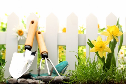 In a well-maintained garden outdoor space, a white picket fence runs across the background, partially obscured by vibrant yellow daffodils and scattered white daisies. In the foreground, there are two small gardening hand tools—a trowel and a cultivator—resting on the soil among freshly sprouted green grass and small plants. The soil appears moist and dark, indicating recent planting activity, and the area is bathed in natural daylight with soft shadows suggesting a bright, partly cloudy day. The garden scene exhibits a neat, organized layout typical of a residential front or back garden, with a combination of grassy patches, flower borders, and soil beds. The overall appearance reflects a space prepared for planting or ongoing gardening work, fitting with the service offerings of local garden maintenance and planting solutions by Landscapers Seven Sisters, serving the SE postcodes. The natural tones of green, yellow, and white create a bright, inviting atmosphere emphasizing healthy growth and outdoor aesthetic care.