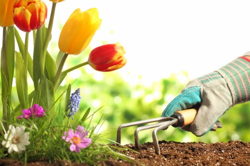 A close-up view of a gardener's gloved hand with a gardening fork, tending to dark, moist soil in a well-maintained garden border. To the left, vibrant tulips in shades of yellow and red, along with various blooming flowers in pink, white, and purple, are visible alongside green foliage. The background shows a softly blurred outdoor environment, indicating a bright, sunny day with natural lighting. The scene highlights elements typical of gardening and landscaping work, with a focus on planting, flower bed maintenance, and outdoor lawn care, reflecting the services offered by Landscapers Seven Sisters in the local area.