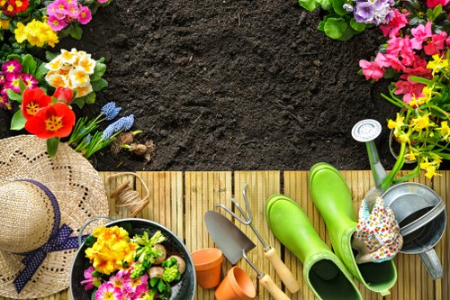 An outdoor garden scene featuring a newly created flower bed bordered with wooden edging, surrounded by vibrant blooming flowers in shades of pink, purple, yellow, and red. The soil in the flower bed appears freshly prepared, with a dark, rich texture. Adjacent to the flower bed, there is a small area of neatly maintained grass, along with garden tools including a watering can, trowel, and pruners resting on a light wooden decking surface. In the foreground, a straw sunhat with a purple ribbon, a pot of colorful flowers, and a pair of bright green rubber gardening boots are visible, suggesting recent planting activity. The background hints at a well-kept outdoor space within the local area, suitable for gardening and landscaping services such as flower bed creation by Landscapers Seven Sisters, serving the SE Postcode area, with natural daylight illuminating the scene.
