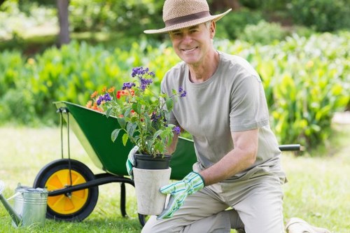A cheerful middle-aged man wearing a straw hat, light grey t-shirt, and gardening gloves kneels on a well-maintained grassy lawn in a garden, holding a container with colorful potted flowers, including purple, red, and orange blooms. Behind him, there is a green garden wheelbarrow with yellow wheels, partially filled with gardening tools, and a metal watering can resting on the grass. The garden features lush, leafy greenery with trees and shrubs in the background, creating a natural and vibrant outdoor setting. Sunlight filters through the foliage, illuminating the scene in soft, natural light typical of a clear day, suggesting a residential garden in the Seven Sisters area. This image highlights outdoor gardening activities supported by professional landscapers specializing in maintaining flower beds, lawns, and garden features for local clients in the vicinity, such as those served by landscaperssevensisters.co.uk.