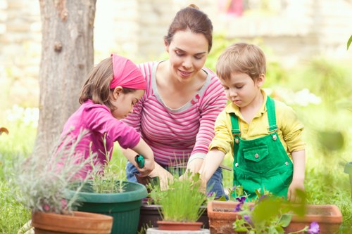 A woman engaged in gardening outdoors in a backyard or garden situated in the Seven Sisters area, surrounded by various potted plants and flowerbeds. The garden features a well-maintained lawn with lush, green grass in the foreground, bordered by flower beds and plant containers made of terracotta and plastic, containing vibrant green foliage and blooming flowers. Behind her, a large tree provides shade, with its trunk and branches visible. The scene shows a natural outdoor environment with a background of blurred garden features and soft sunlight filtering through the trees. The woman is focused on tending to the plants, highlighting outdoor lawn and garden maintenance activities typical of professional services offered by Landscapers Seven Sisters, emphasizing landscaped outdoor spaces suited for family activities and gardening enthusiasts in the local area.
