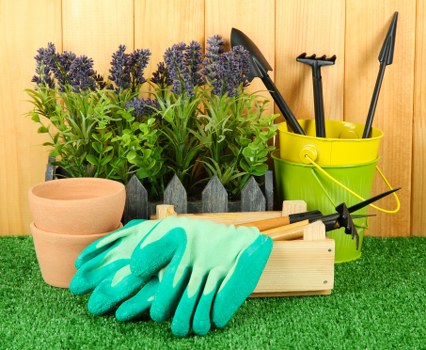The image displays a neatly arranged garden storage area with various gardening tools and accessories. In the foreground, there are a pair of bright teal gardening gloves laid over a small wooden box, which holds a black garden tool with a curved blade. To the left, three terracotta plant pots of different sizes are stacked together, positioned on a well-maintained artificial grass surface that covers the ground. Behind the pots, a row of purple flowering lavender plants with green foliage stands upright, creating a colourful and fragrant backdrop. Towards the right side, a yellow plastic bucket contains several gardening hand tools with black handles, including a trowel, a fork, and a hand rake. The background features a vertical wooden fence, typical of garden enclosures, which provides a warm and natural setting for outdoor gardening activities. The scene was captured in bright daylight, suggesting clear weather, with natural light highlighting the vibrant plant colours and the textured surfaces of the gardening accessories, subtly reflecting the lawn care and gardening services offered by landscapers across Seven Sisters and the surrounding area.