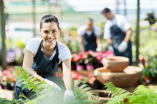 A cheerful female gardener wearing a white t-shirt and striped apron is tending to lush green ferns in a bright, spacious greenhouse or garden centre environment. Behind her, two other gardeners are visible, one in a dark apron and the other in a white shirt with a dark vest, working among vibrant pink flowers and potted plants arranged on tiered wooden display stands. The setting features a well-maintained outdoor garden area with dense grass, neatly edged flower beds, and a mix of leafy shrubs and small trees under natural daylight. The background shows a greenhouse structure with glass panels allowing natural light to illuminate the scene, creating a warm, inviting atmosphere suited for professional gardening and garden maintenance services in the Seven Sisters or nearby postcode area. The garden layout emphasizes a combination of cultivated planting beds, gravel pathways, and plant care activities, illustrating professional outdoor landscaping and gardening expertise in a vibrant, natural setting.